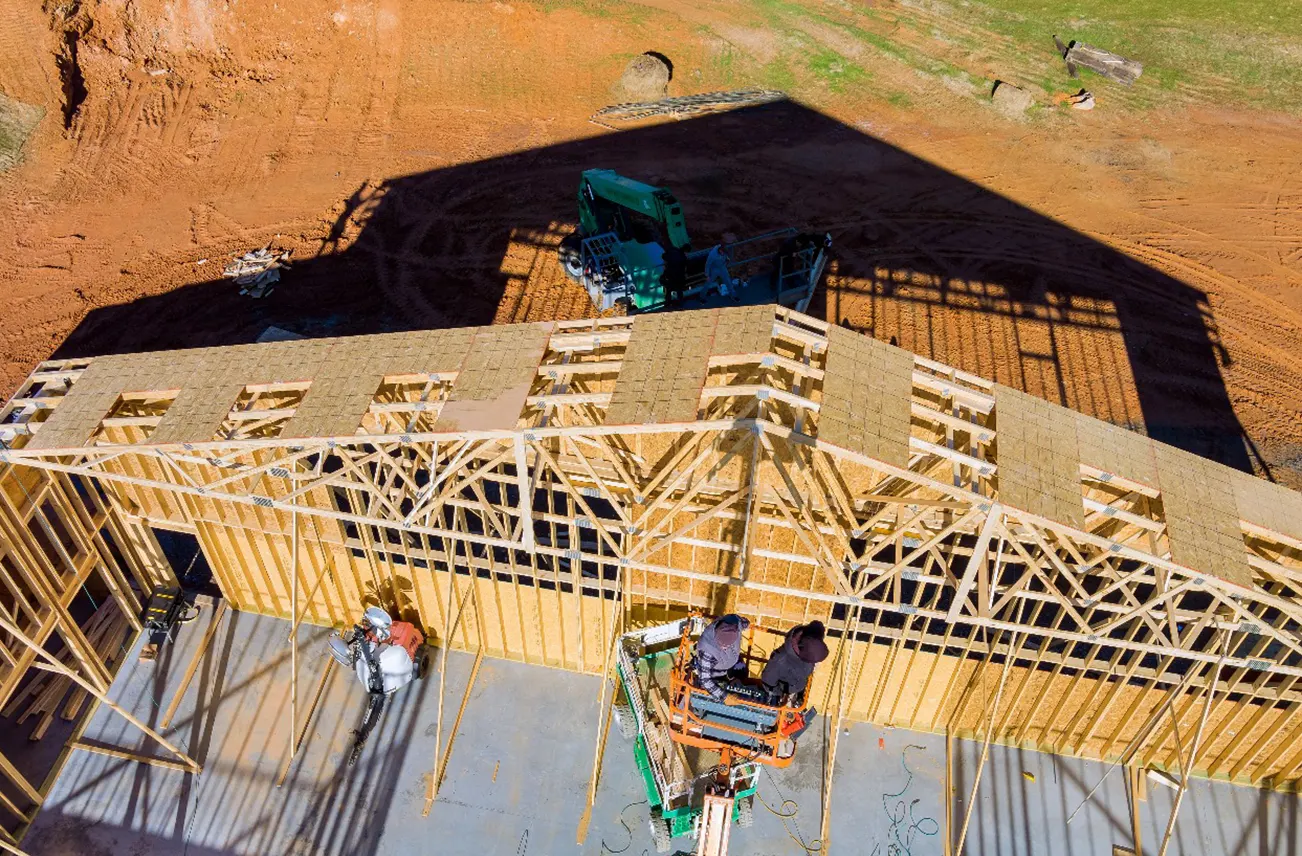 Overhead view of a home under construction with wooden framing and workers on-site, highlighting structural integrity. Sunstate Roofs in Central Florida specializes in residential services, including Roof Replacement cost analysis, relief services, tape repairs, and nearby roof replacement options.