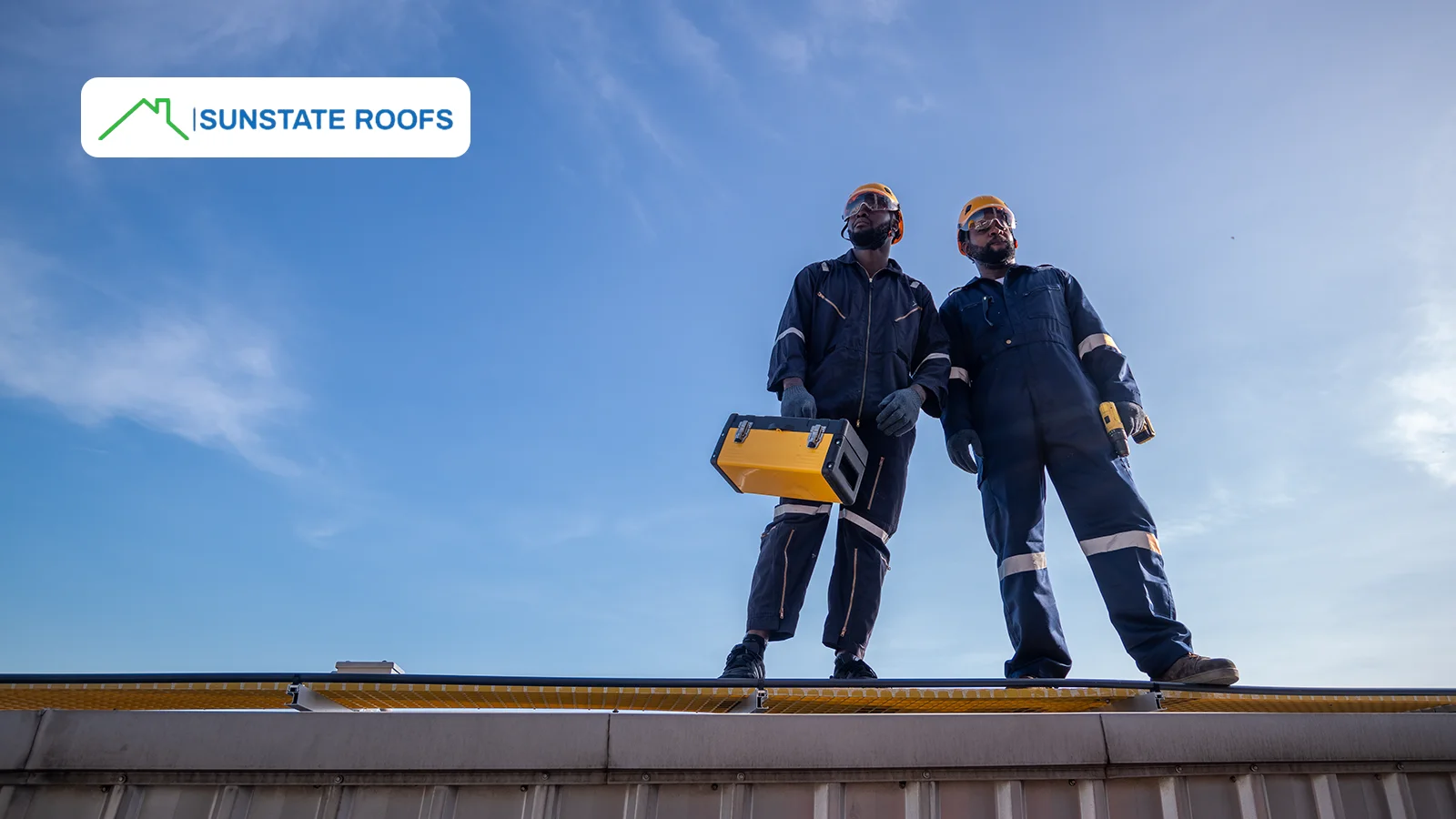 Two professional roofers wearing safety gear standing on a commercial rooftop, inspecting the structure under a clear blue sky.